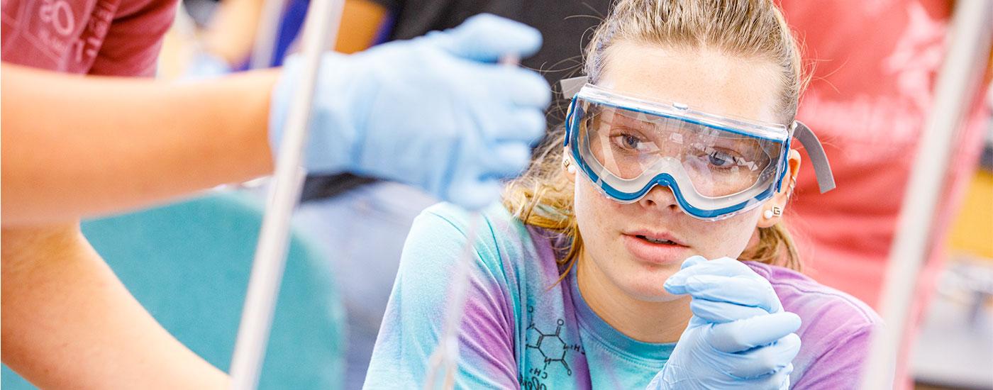 A female Frostburg State student wearing safety goggles observes another student put a liquid in a beaker during a chemistry experiment